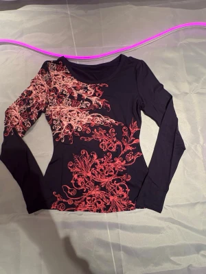 Svart långärmad topp med rosa mönster - Snygg svart långärmad topp med unikt rosa blommigt mönster över ena axeln och sidan. Figurnära passform och rund halsringning. Perfekt för dig som vill sticka ut med coola detaljer. Aldrig använd 