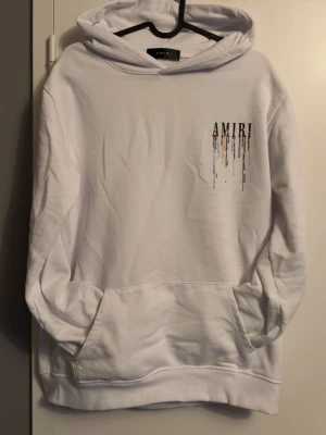 Vit hoodie från Amiri med färgstänk - Säljer en vit hoodie från Amiri med stor huva och känguruficka. På bröstet och ryggen står det AMIRI med färgstänk i svart, orange, blått och lila som rinner ner. Hoodien har en avslappnad passform och ribbade muddar vid ärmar och nederkant. Perfekt för dig som gillar streetwear och unika detaljer.
