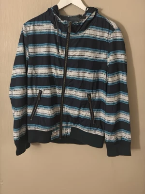 Y2K Randig vindjacka från Levi's - Snygg vindjacka från Levi's med blå, vita och turkosa horisontella ränder. Jackan har huva, dragkedja framtill och två fickor med dragkedja. Perfekt för dig som vill ha en lätt och stilren jacka med coolt mönster.