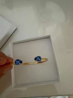 Caroline Svedbom  - Snyggt och stilrent armband i guldfärgad metall med två stora blå glittrande stenar i ändarna. Armbandet är öppet och har en minimalistisk design som känns både trendig och elegant. Perfekt accessoar för att lyfta din outfit.