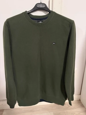 Olivgrön tröja från lionette - Snygg olivgrön tröja från lionette med rund halsringning och diskret logga på bröstet. Tröjan har långa ärmar och är tillverkad i ett mjukt, tunt stickat material som passar perfekt till lager på lager. Enkel och stilren design.