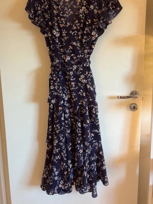 Marinblå blommig långklänning - Snygg marinblå långklänning med vitt och beige blommönster. Klänningen har v-ringning, volangdetaljer och ett knytband i midjan. Tillverkad i ett lätt och luftigt material, perfekt för varma dagar. Ärmarna är korta och fladdriga för en avslappnad look.