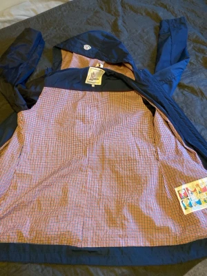 Blå vindjacka från Moncler Grenoble - Snygg blå vindjacka från Moncler Grenoble med rutigt innerfoder i rött, vitt och blått. Jackan har huva och klassisk Moncler-logga på insidan. Materialet är tunt och smidigt, perfekt för blåsiga dagar. Stängs med dragkedja och har en cool retrovibe. Passar både herr och dam. dam storlek S eller M. Annas 2. Det finns några skador men går att sy det syns inte när man har på sig den. Skriv om ni är intresserade eller diskutera priset 