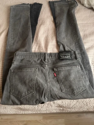 Grå Levi's jeans med raka ben - Säljer ett par grå Levi's jeans med raka ben och klassiska bakfickor. Jeansen har en vintage look med slitna detaljer och råa benslut. Tillverkade i bomull med den ikoniska Levi's-lappen bak i midjan och röd etikett på fickan.