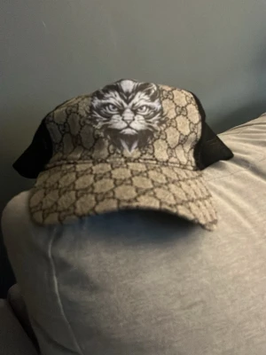 Gucci keps med kattmotiv - Säljer en snygg Gucci keps med klassiskt GG-mönster i beige och brunt, svart mesh baktill och ett stort kattmotiv framtill. Kepsen har justerbar rem bak och är tillverkad i en mix av bomull och mesh. Perfekt för dig som vill sticka ut med en unik accessoar.