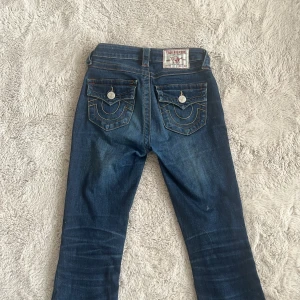 True Religion mörkblå bootcut jeans - Säljer ett par mörkblå bootcut jeans från True Religion med ikoniska fickor och silverfärgade knappar bak. Jeansen har kontrastsömmar och klassisk femficksdesign. Perfekta för dig som gillar streetwear och vill sticka ut med detaljer.