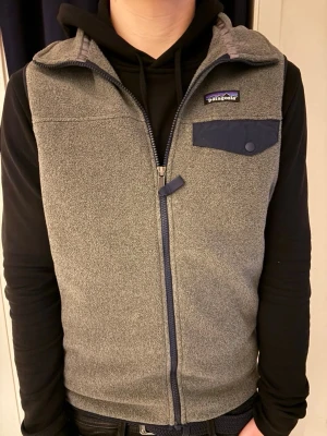 Grå fleeceväst från Patagonia - Säljer en grå fleeceväst från Patagonia med hel dragkedja framtill, hög krage och tre fickor med blå detaljer. Västen har en bröstficka med knapp och två sidofickor med dragkedja. Perfekt för lager-på-lager och riktigt skön att bära.