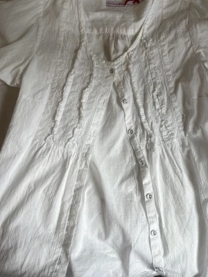 Vit blus med knappar och volanger - Söt vit blus från Urban Girl med korta ärmar, knappar framtill och dekorativa volanger längs bröstet. Blusen har en lätt rynkad midja och tunna vertikala ränder i tyget för extra struktur. Perfekt till jeans eller kjol.