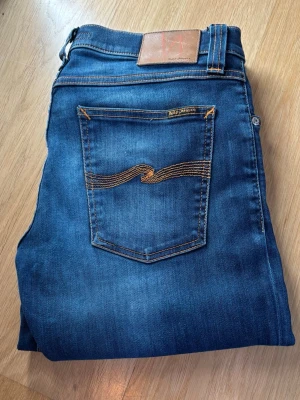 Blå skinny jeans från Nudie Jeans - Snygga blå skinny jeans från Nudie Jeans med klassisk femficksmodell och kontrastsömmar. Jeansen har en cool tvärt med lätt slitning och bruna detaljer på bakfickan. Perfekta för en trendig och avslappnad stil. Hör av dig vid funderingar 🔥👍🔥👍