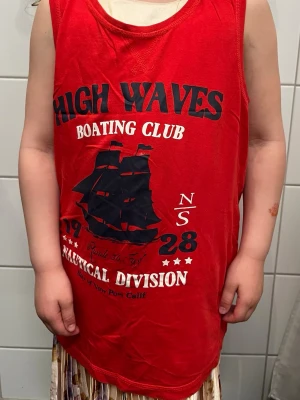Röd ärmlös linnetopp med tryck - Cool röd linnetopp med svart och vitt tryck framtill, texten 'High Waves Boating Club' och en stor segelbåt. Toppen är ärmlös och har rund halsringning. Perfekt för en avslappnad och sportig look.