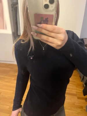 Svart långärmad tröja från Gant - Snygg svart långärmad tröja från Gant med liten broderad logga på bröstet. Tröjan har en slim fit passform och är tillverkad i mjukt material som sitter skönt på kroppen. Perfekt att matcha med jeans eller kjol för en clean look.