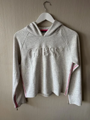 Ljusgrå hoodie från Guess - Snygg ljusgrå hoodie från Guess med broderad logga framtill. Kolla gärna i bild 4. Bredden: 45cm Längden: 50cm. Stor inga storlek men jag har skrivit mätningar. 