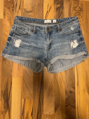 Blå denimshorts med slitningar - Säljer ett par klassiska blå jeansshorts med slitna detaljer och upprullade benslut. Shortsen har fem fickor och stängs med knapp och dragkedja. Perfekta för sommaren och ger en avslappnad vibe.