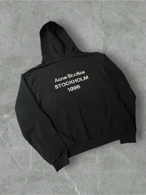 Svart hoodie Acne Studios | L/XL - Svart hoodie från Acne Studios med tryck i vitt på ryggen där det står 'Acne Studios STOCKHOLM 1996'. Klassisk passform med huva och ribbade muddar. Perfekt för dig som gillar minimalistisk och stilren streetwear.