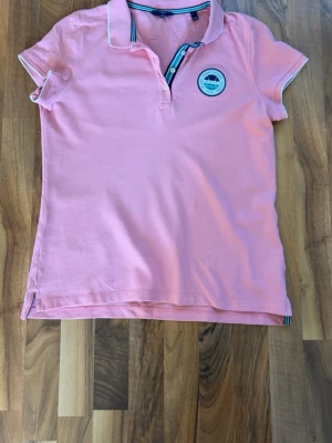 Rosa pikétröja från Gant - Snygg rosa pikétröja från Gant med vit logga broderad på ryggen och ett märke på bröstet. Tröjan har korta ärmar, krage med knappar och tunna vita detaljer längs kragen och ärmsluten. Tillverkad i mjuk bomull för en bekväm känsla.