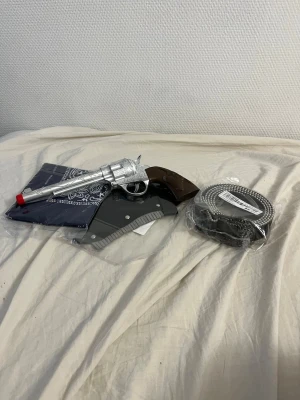 Cowboy maskeradset med leksakspistol - Cowboy maskeradset med silverfärgad leksakspistol, svart bälte, blå bandana och hölster. Perfekt för dig som vill köra westernstil på maskerad eller cosplay. Pistolen har brunt handtag och röd topp. Allt är i plast och tyg.