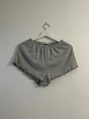 Grå ribbade shorts med volangkant - Mysiga grå shorts med ribbad struktur och volangkant nertill. Shortsen har elastisk midja och är perfekta för avslappnade dagar. De är tillverkade i mjukt material och har en enkel, stilren look.