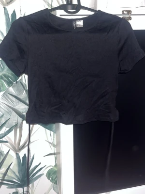 Svart croppad topp med snörning - Snygg svart croppad topp från H&M med korta ärmar och snören i ryggen för en trendig look. Tillverkad i mjuk bomull som känns skön mot huden. Perfekt att matcha med jeans eller kjol.