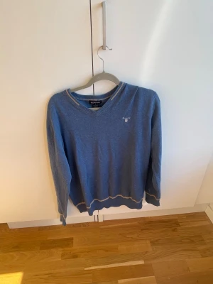 Blå tröja från Gant med V-ringning - Blå tröja från Gant med V-ringning och diskret logga broderad på bröstet. Tröjan har kontrasterande detaljer vid hals, ärmslut och nederkant. Perfekt för en avslappnad men ändå stilren look.