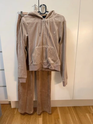 Beige velourset från Juicy Couture - Mysigt loungewear-set i beige velour från Juicy Couture. Setet består av en zip-hoodie med fickor och dragsko i huvan som är storlek M samt matchande byxor med bootcut ben storlek S. Perfekt för chill dagar hemma eller när du vill vara extra bekväm.