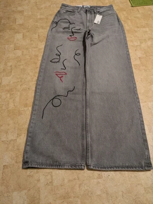 Gråa vida jeans med ansiktsbrodyr - Unika grå jeans med bred passform och konstnärlig brodyr av ansikten i svart och rött på ena benet. Jeansen har hög midja, klassiska fickor och är tillverkade i denim. Perfekta för dig som vill sticka ut med en kreativ och trendig look.