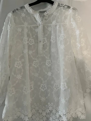 Vit spetsblus med blommönster - Romantisk vit blus i transparent spets med broderade blommor och dekorativa knappar framtill. Blusen har lång ärm och rund halsringning med spetskant. Perfekt för dig som gillar en feminin och trendig stil. Materialet är lätt och luftig bomull. Aldrig använd men råkade dra av taggen.