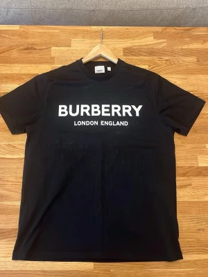Svart Burberry t-shirt med logga - Snygg svart t-shirt från Burberry med stor vit logga 'BURBERRY LONDON ENGLAND' på bröstet. Klassisk rund hals och korta ärmar. Tillverkad i mjuk bomull som känns skön mot huden. Perfekt för dig som gillar stilrena och exklusiva plagg.