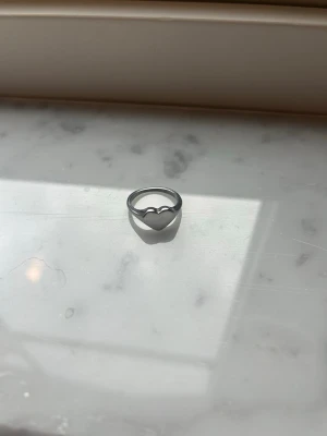 Silverring med hjärtformad topp - Snygg silverfärgad ring med en hjärtformad topp. Ringen har en enkel och stilren design som passar perfekt för dig som gillar minimalistiska smycken. Tillverkad i metall med slät yta och diskret gravyr på insidan. Skriv vid frågor om storlek, så kan jag mäta!