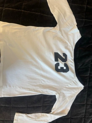 Vit långärmad t-shirt  - Snygg vit långärmad t-shirt från LA GEAR med svart nummer 23 tryckt. Tröjan har svarta detaljer vid kragen och är tillverkad i mjuk bomull, perfekt för en avslappnad och sportig stil.