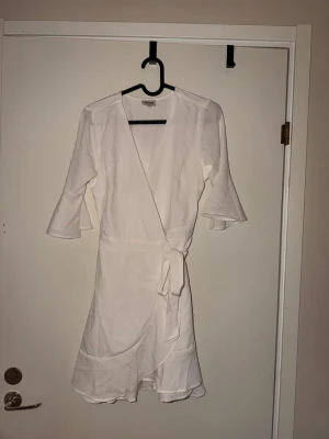 Vit omlottklänning med volangärm skolavslutningsklänning - Snygg vit omlottklänning med trekvartsärm och volangdetaljer. Klänningen har knyt i midjan och lätt v-ringad hals. Tillverkad i ett tunt, luftigt material. Perfekt till skolavslutningen!