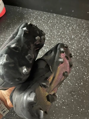 Svarta Umbro fotbollsskor - Säljer ett par svarta fotbollsskor från Umbro med klassisk snörning och stora vita Umbro-loggor på sidorna. Skorna har fasta dobbar för bra grepp på planen och är tillverkade i ett slitstarkt syntetmaterial. Perfekta för dig som vill ha stilrena och funktionella skor.