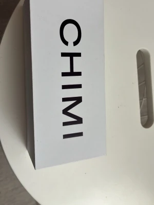 Gråa Chimi-Glasögon - Säljer ett par stilrena solglasögon från CHIMI. Kommer i originalförpackning med ett vitt, strukturerat fodral där CHIMI-loggan är präglad. Perfekt accessoar för dig som vill ha en clean och modern look.