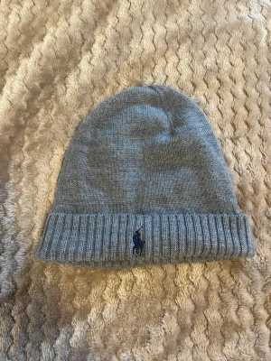 Grå merinoullsmössa Polo Ralph Lauren - Snygg grå mössa från Polo Ralph Lauren i mjuk och tvättbar merinoull. Klassisk ribbad kant med broderad mörkblå logga framtill. Perfekt accessoar för kyliga dagar och enkel att matcha med olika stilar.