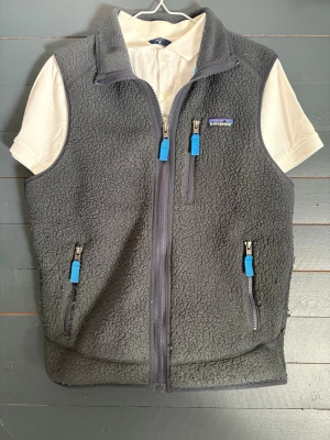 Grå fleeceväst från Patagonia - Mysig grå fleeceväst från Patagonia med blå detaljer på dragkedjorna och tre fickor med dragkedja. Västen har hög krage och klassisk Patagonia-logga på bröstet. Perfekt för lager-på-lager och outdoor-vibe.