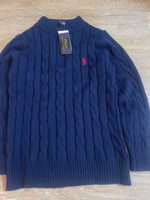 NEW   tröja Polo Ralph Lauren Storlek M - Mörkblå kabelstickad tröja från Polo Ralph Lauren med halv dragkedja och ribbade muddar. Klassisk röd logga broderad på bröstet. Perfekt för dig som gillar stilren och tidlös design. Materialet känns mjukt och varmt.