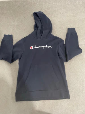 Mörkblå Champion hoodie med huva - Snygg mörkblå hoodie från Champion med stor logga broderad på bröstet. Klassisk känguruficka framtill och skön huva. Tillverkad i mjukt bomullsmaterial som passar perfekt till en avslappnad stil.