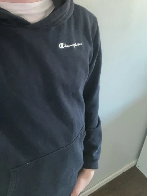 Mörkblå hoodie från Champion - Snygg mörkblå hoodie från Champion med klassisk logga broderad på bröstet. Hoodien har en stor känguruficka framtill och en skön huva. Perfekt för dig som gillar streetwear och vill ha en enkel men stilren look. Den är lite smutsig. Den hade ett champion märke på handleden på tröjan som har tagits bort det syns lite. Jag är villig att kunna sänka priset om nån vill köpa. Jag säljer den här för jag vill ha pengar.