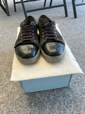 Svarta låga sneakers med lackdetaljer från Lanvin - Snygga svarta sneakers med mockaliknande material på sidorna och glansiga lackdetaljer på tån. Skorna har svarta snören och silvriga öljetter. Klassisk vit sula som ger en clean look. Perfekta för dig som vill ha en stilren men ändå unik sneaker. Skorna köptes på Plick för ungefär ett år sedan och är i ganska dåligt skick men går ändå att använda är öppen för byten men då måste det vara ett riktigt bra byte🙌 Hör av dig vid frågor🌟👍