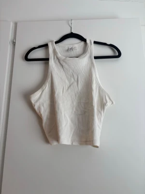 Vit ribbad croppad linnetopp - Säljer en vit croppad linnetopp, ribbad. Toppen är ärmlös med breda axelband och har en sportig, relaxed fit. Perfekt till sommaren och enkel att matcha med olika stilar.