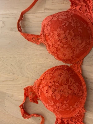 Orange spets-bh med vaddering - Snygg orange bh med vadderade kupor och blommig spets över hela framsidan. Justerbara axelband och hakknäppning bak. Bh:n har en feminin och ungdomlig look med fina spetsdetaljer. Perfekt för dig som vill ha något färgglatt och bekvämt. Använd 1 gång tvättad. Storlek C