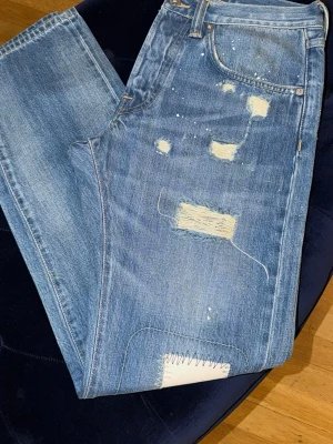 Blå distressed jeans från Edwin - Säljer ett par blå jeans från Edwin med slitna detaljer, lagningar och färgstänk för en riktigt cool och avslappnad look. Klassisk femficksmodell i denim med patch på bakfickan och läderpatch i midjan. Perfekta för dig som gillar streetwear och unik stil.