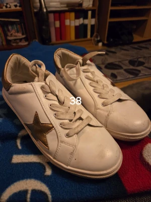 Vita sneakers med guldstjärna - Snygga vita sneakers med en metallic guldstjärna på sidan och gulddetaljer vid hälen. Klassisk låg modell med vita skosnören och rund tå. Perfekta för dig som vill ha en clean look med en twist.