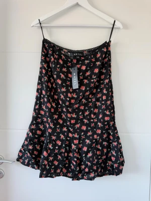 Svart blommig kjol från Motel Rocks - Söt svart kjol från Motel med  små röda blommor över hela tyget. Kjolen har en lätt utsvängd nederdel och är tillverkad i ett mjukt, glansigt material som ger en skön känsla. Perfekt för sommarens alla tillfällen!