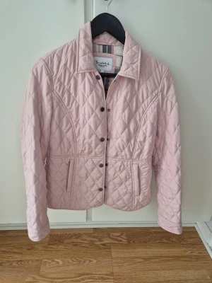 Ljusrosa quiltad jacka - Snygg ljusrosa quiltad jacka från Bondelid med tryckknappar och dragkedja framtill. Jackan har lång ärm och figurnära passform, perfekt för vår och höst. Rutigt innerfoder. Väldigt feminin och stilren. 