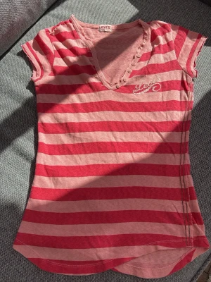 Y2K Levi’s Randig rosa t-shirt  - Säljer en kortärmad t-shirt från Levi's i rosa och rött med breda ränder. T-shirten har v-ringning med dekorativa knappar och ett broderat Levi's-tryck på bröstet. Tillverkad i mjuk bomull och har en normal passform.