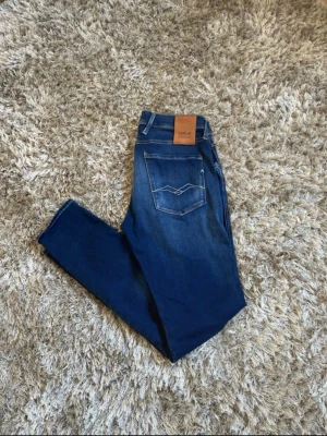 Blå jeans från Replay med stretch - Säljer ett par snygga blå jeans från Replay med klassisk femficksmodell och vita kontrastsömmar. Jeansen har en skön stretch, normal midja och en snygg tvätt med slitningar framtill. Perfekta till en avslappnad stil.