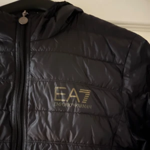 Svart Emporio Armani Jacka - Snygg svart pufferjacka från Emporio Armani EA7 med huva och dragkedja framtill. Jackan har quiltad design och logga på bröstet. Perfekt för kalla dagar och ger en sportig look. Tillverkad i lätt, glansigt material som håller värmen.