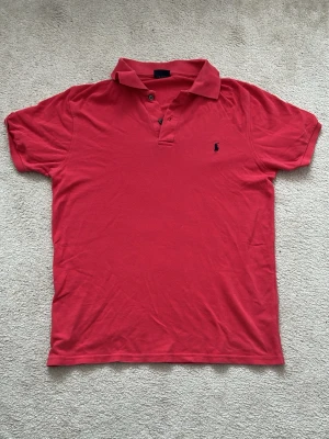 Röd pikétröja från Polo Ralph Lauren - Klassisk röd pikétröja från Polo Ralph Lauren med korta ärmar och blå broderad logga på bröstet. Tröjan har krage och knappar vid halsen. Perfekt till jeans eller shorts för en avslappnad men snygg look. L men passar även M