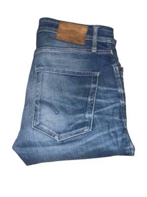 Blå jeans från Jack & Jones - Säljer ett par klassiska blå jeans från Jack & Jones med snygg tvätt och slitningar. Jeansen har fem fickor, normal midja och raka ben. Tillverkade i slitstark denim med läderpatch bak i midjan. Perfekta för dig som gillar en avslappnad men stilren look.