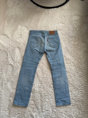 Ljusblå raka jeans från Levi's 501 - Säljer ett par klassiska ljusblå jeans från Levi's med rak passform och fem fickor. Jeansen har den ikoniska läderpatchen bak i midjan och röda Levi's-taggen på bakfickan. Tillverkade i slitstark denim, perfekta för en avslappnad och trendig look.